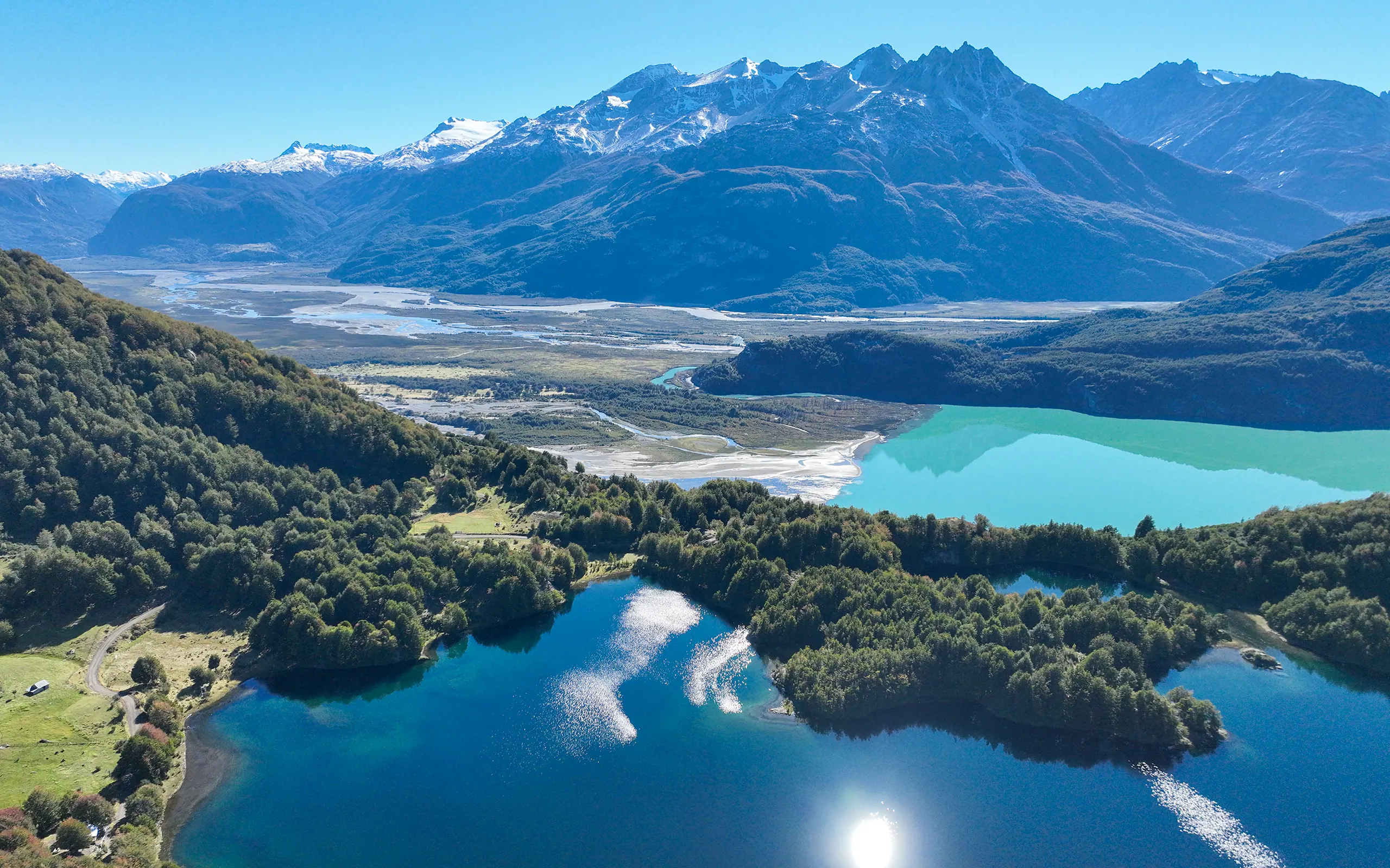 Vista aérea de un valle de Aysén: lagunas azules entre bosque nativo, un río trenzado bajando por el valle, un lago turquesa a un costado y la cordillera nevada al fondo