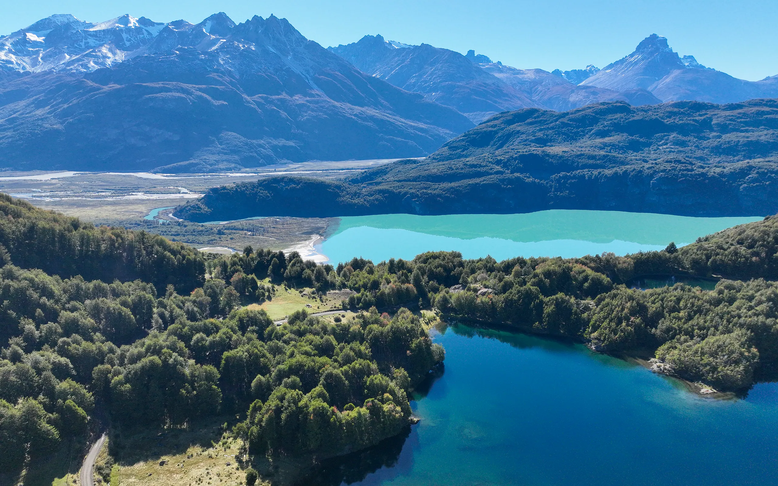 Vista aérea de un lago turquesa de origen glaciar y un lago azul rodeados de bosque, con la cordillera nevada al fondo